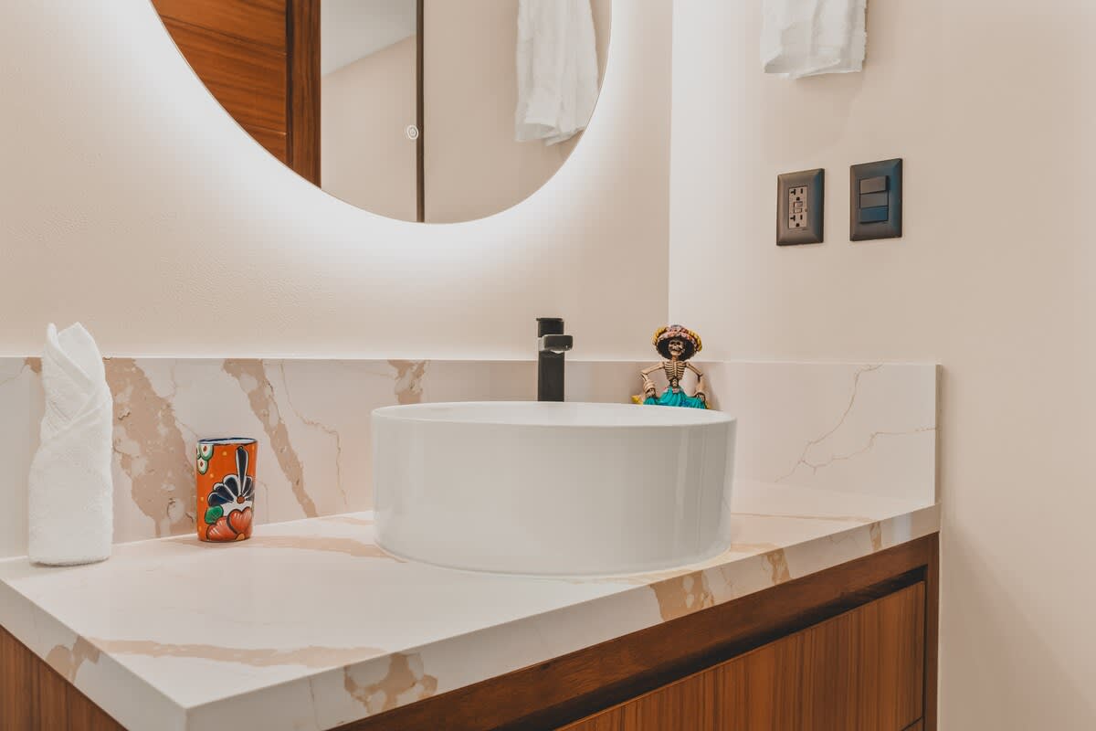 A modern bathroom vanity features a white vessel sink, a black faucet, and a light marble countertop with brown veining, accented by a colorful ceramic cup and a decorative skeleton figurine.