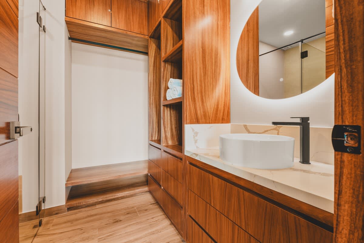 Spacious walk-in closet with rich wooden cabinetry and shelving, leading into a modern bathroom vanity area with a round illuminated mirror and a vessel sink.