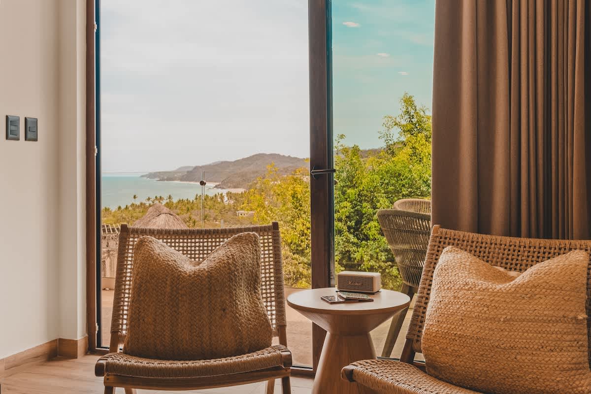 Cozy seating area with two woven chairs and a small round side table, positioned by a large glass door that opens to a lush green landscape and a distant ocean view.