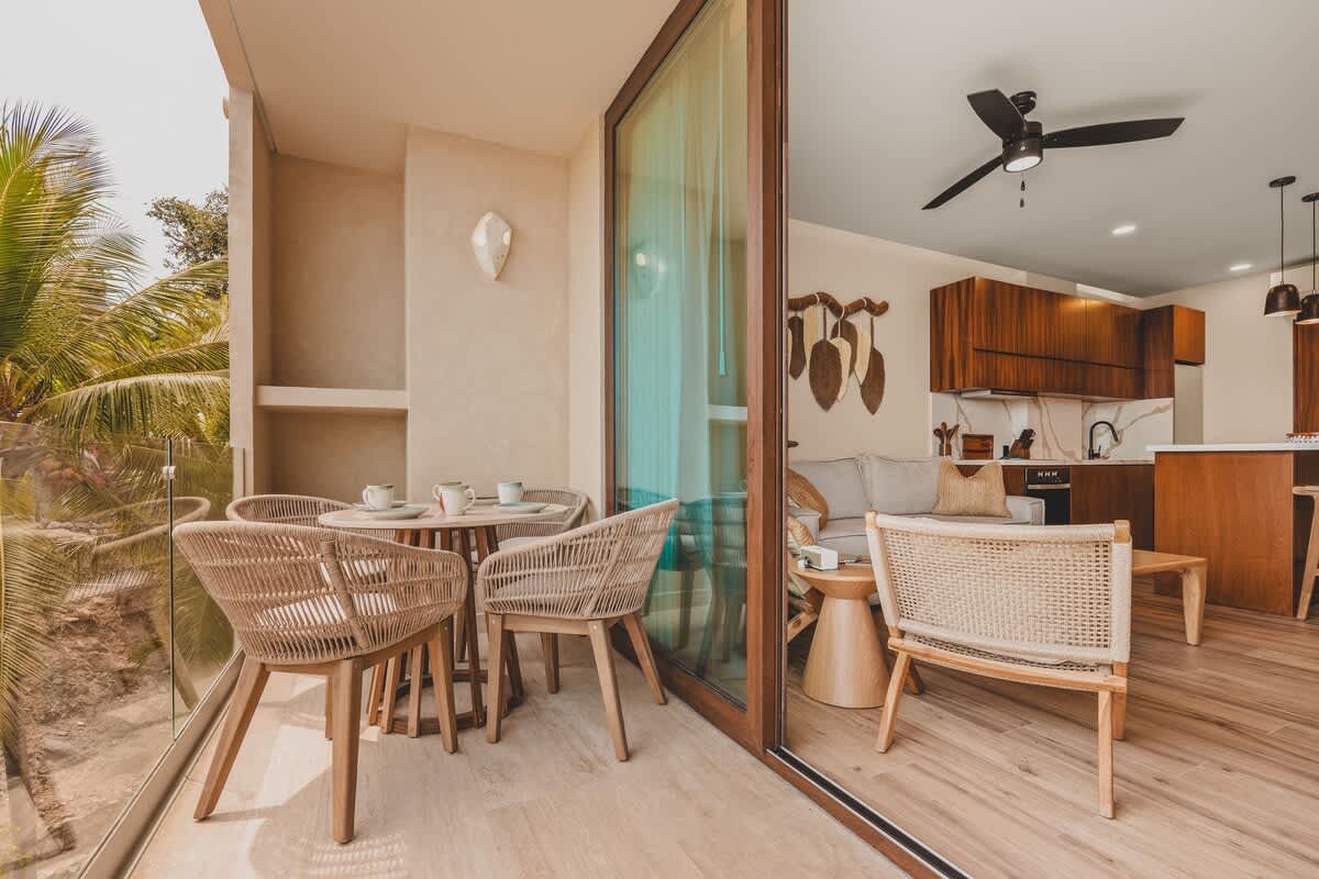 A private balcony with a round dining table and four woven chairs, separated by a large glass sliding door from an interior living space, offering a glimpse of lush greenery outside.