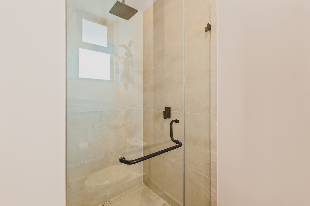 A modern walk-in shower features light-colored tiled walls, a square rain showerhead, and a clear glass enclosure with a black handle, offering a clean and minimalist design.