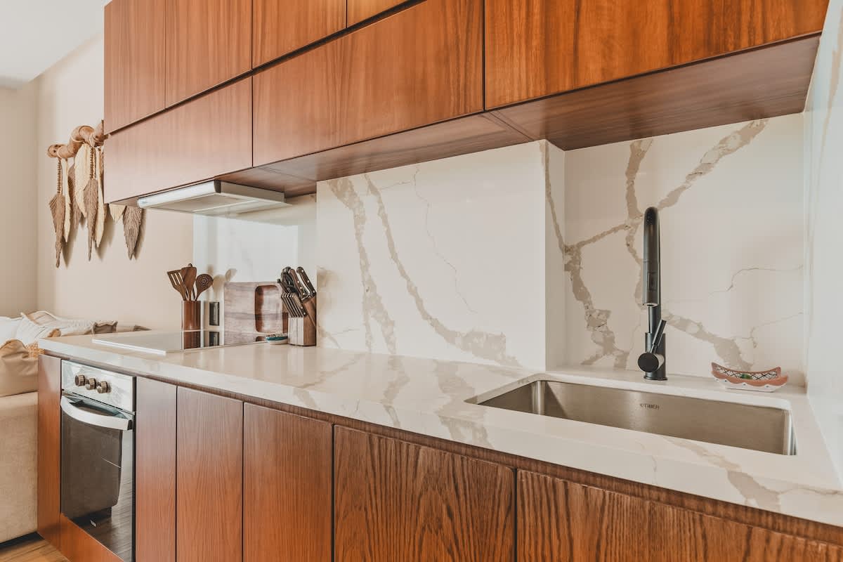 A modern kitchen counter with a stainless steel sink and a matte black gooseneck faucet, set against a white quartz backsplash with prominent grey veining. Dark wood cabinetry is visible above and below the counter.