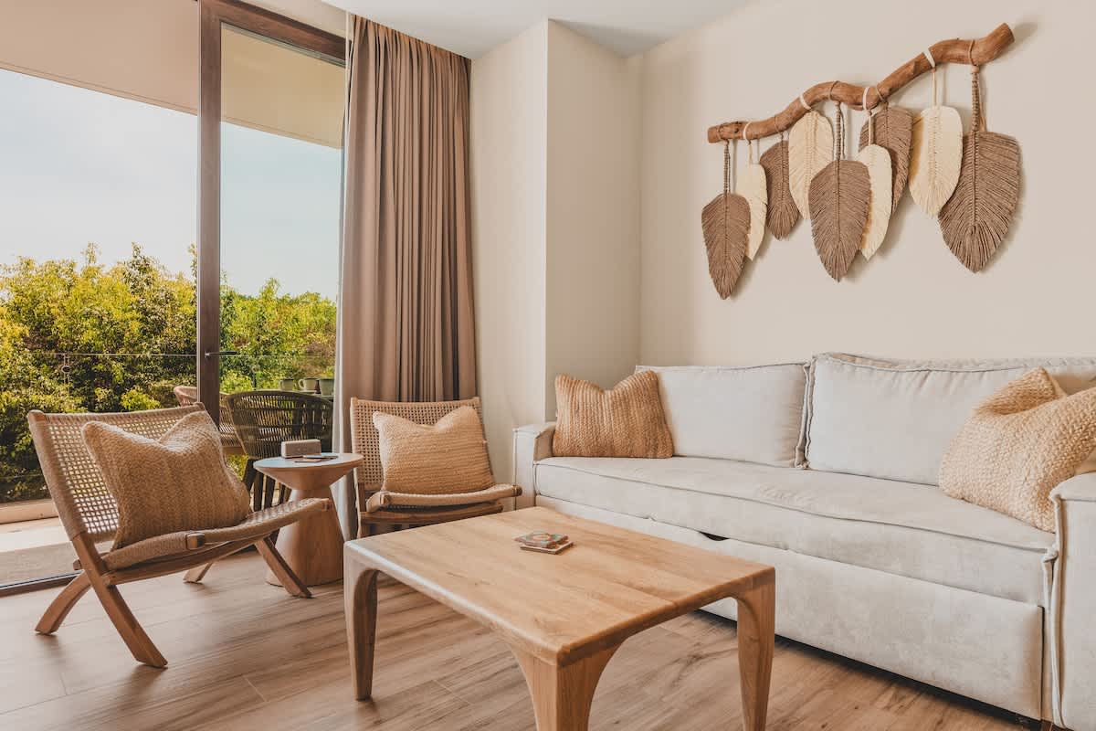 A cozy living room with a light-colored sofa, a wooden coffee table, and two woven armchairs. A large sliding glass door opens to a balcony with lush greenery, and a decorative wall hanging with feather motifs adds a bohemian touch.