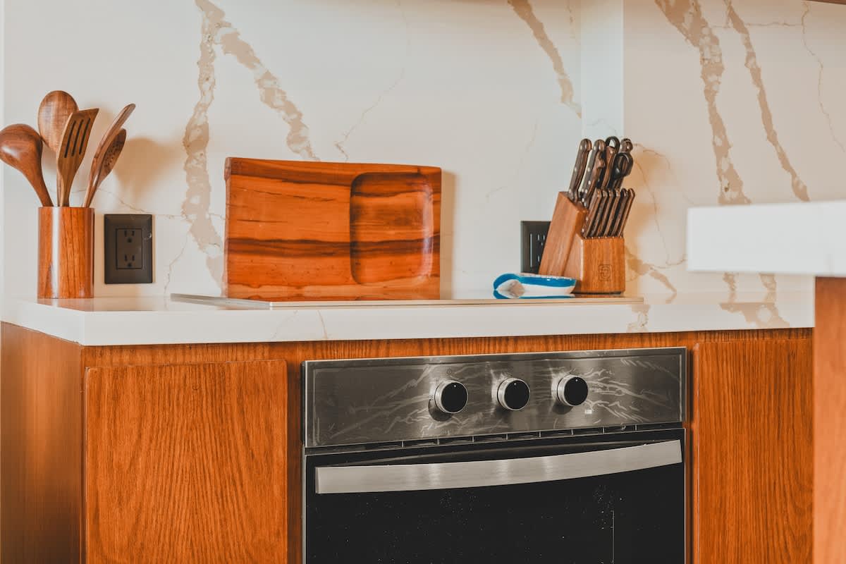 Modern kitchen counter with a white marble backsplash featuring grey veining, a built-in stainless steel oven, and wooden cabinetry, accessorized with wooden cutting boards and utensils.