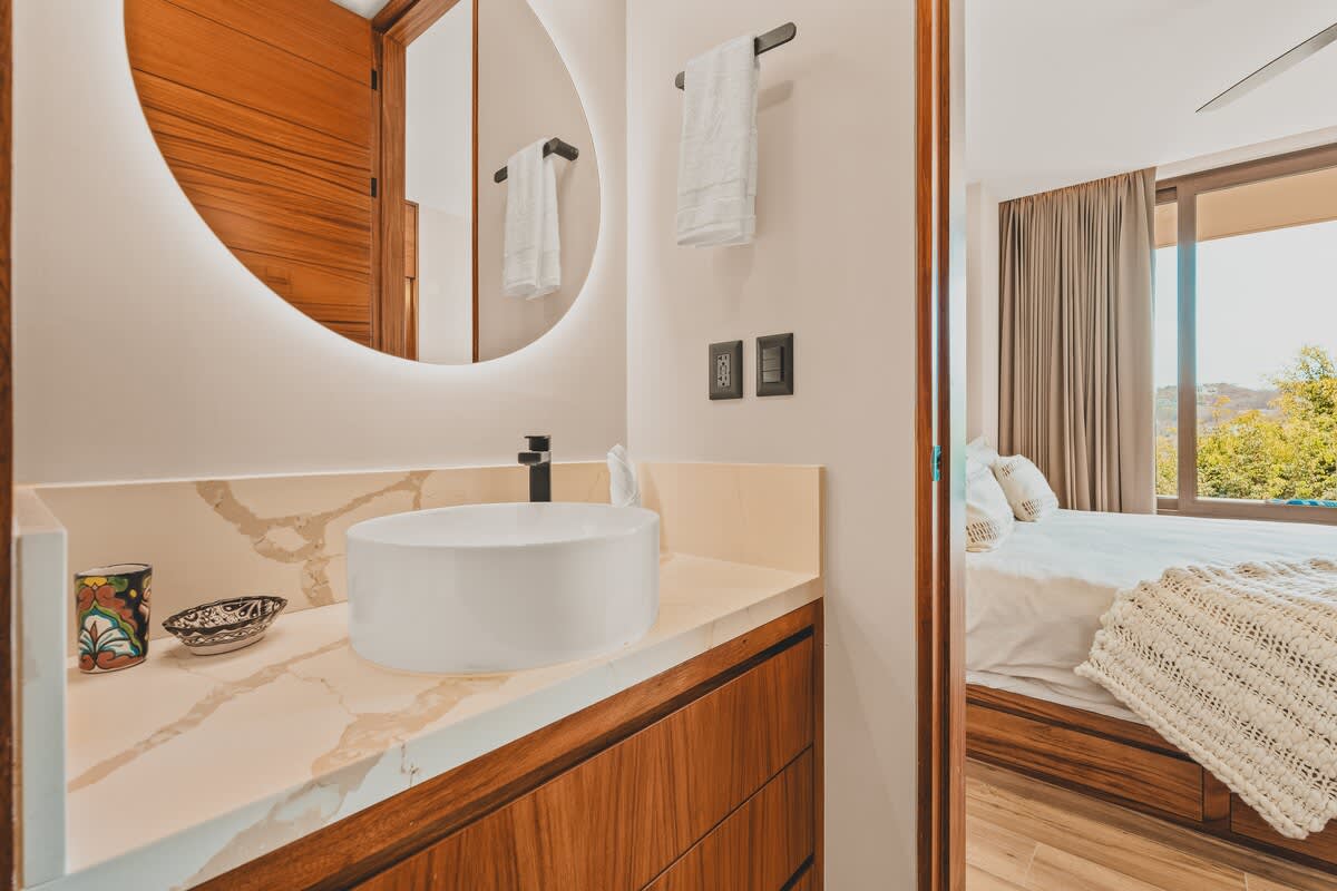 View of the bathroom vanity with a vessel sink and illuminated mirror, showing the open doorway leading into a bedroom with a bed visible in the background.