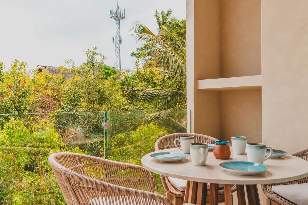 Outdoor balcony with a round wicker dining table and chairs, set against a backdrop of dense tropical foliage and a distant cell tower, offering a casual al fresco dining experience.