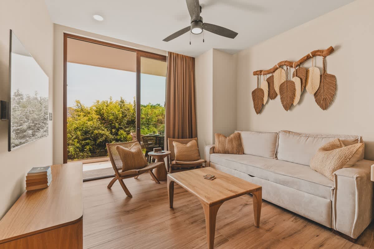Bright living room featuring a light-colored sofa, a wooden coffee table, and two woven chairs, with a large sliding glass door opening to a balcony overlooking lush greenery.