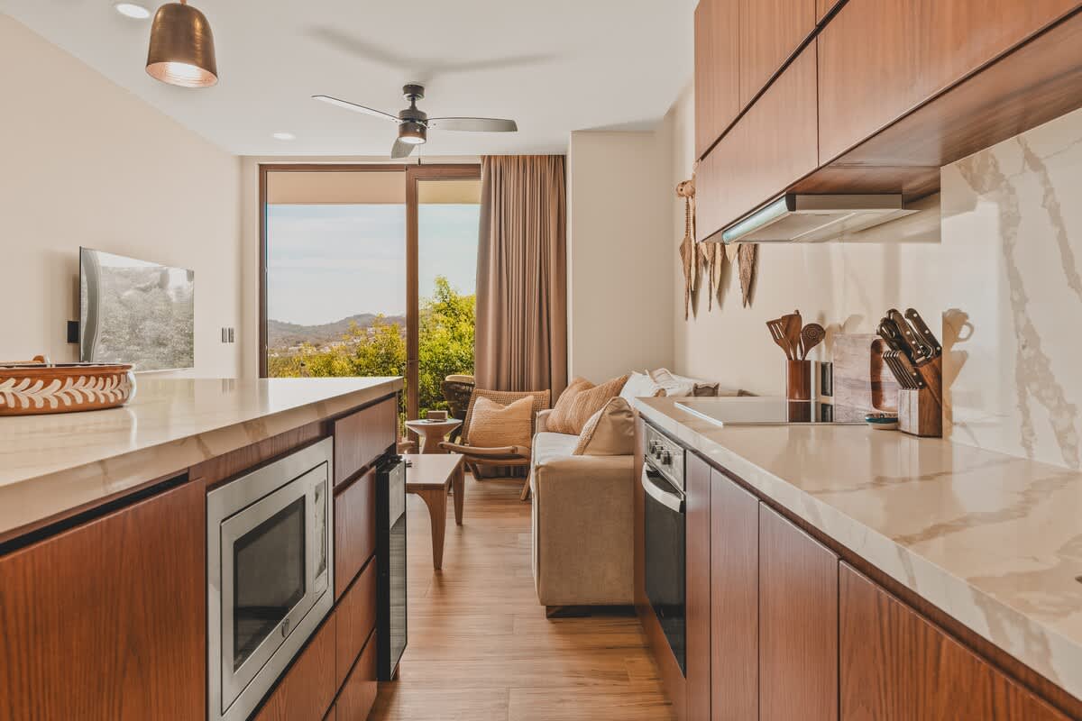 A sleek kitchen with dark wood cabinetry and white quartz countertops, featuring an integrated microwave below the counter and an oven. The view extends to an open living area with a sofa and a balcony overlooking lush green trees and distant hills.