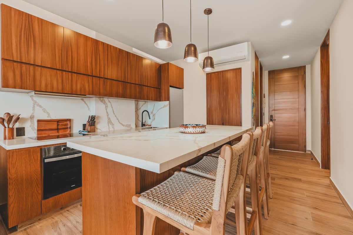 An expansive view of the open-concept kitchen and dining area, featuring a long white quartz island with four woven bar stools. Dark wood cabinetry lines the walls, and three bronze pendant lights hang above the island, leading to a hallway on the right.