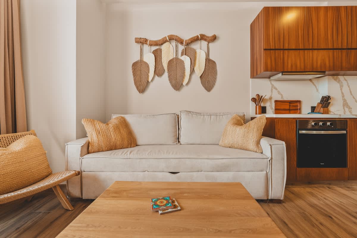 Comfortable living room with a light-colored sofa and throw pillows, a wooden coffee table with coasters, and a decorative wall hanging, with the modern kitchen visible in the background.