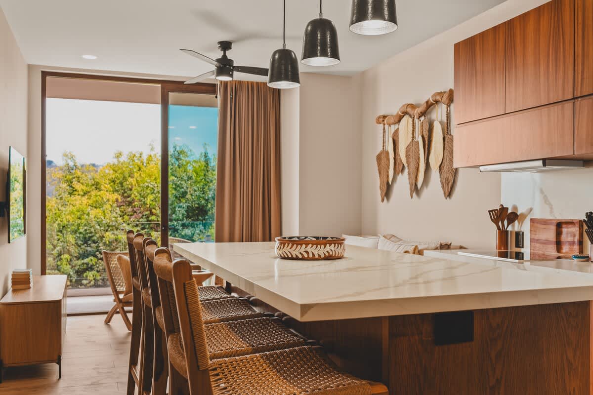 Open-concept kitchen and living area featuring a long kitchen island with bar seating, dark wooden cabinetry, and a decorative wall hanging, with a view towards the outdoor landscape through a large glass door.