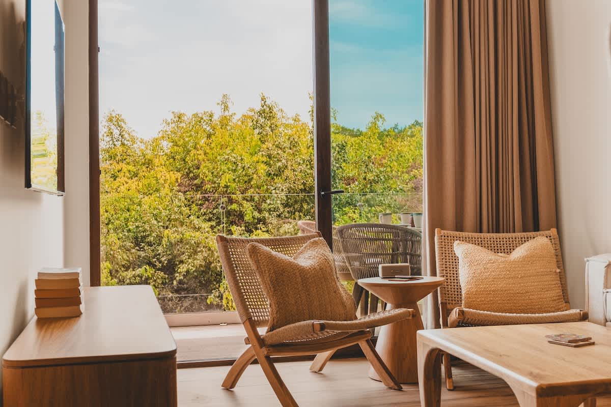 A bright living area features two woven armchairs with natural-toned cushions, a wooden coffee table, and a console, all facing a large glass door leading to a balcony with lush green views.