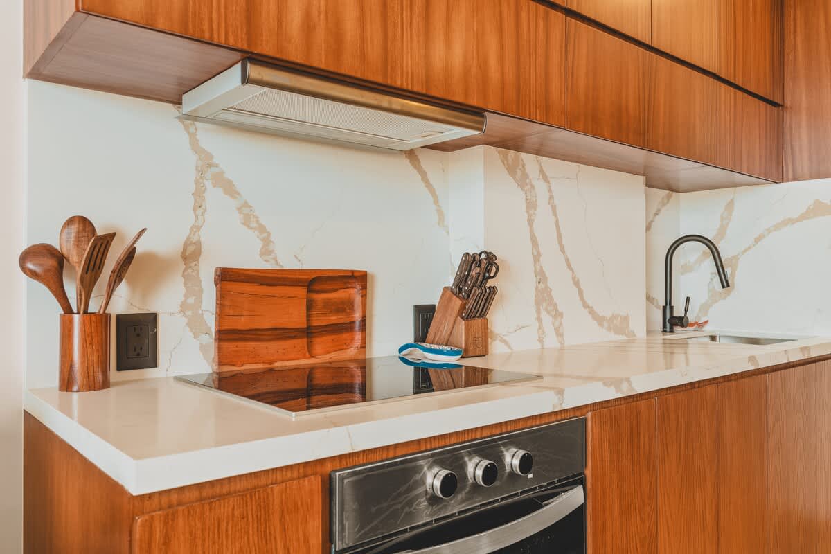 A contemporary kitchen featuring a built-in electric stovetop and oven, with a stainless steel range hood mounted above. The white quartz countertop and backsplash display prominent grey veining, complemented by dark wood cabinetry and wooden kitchen utensils.