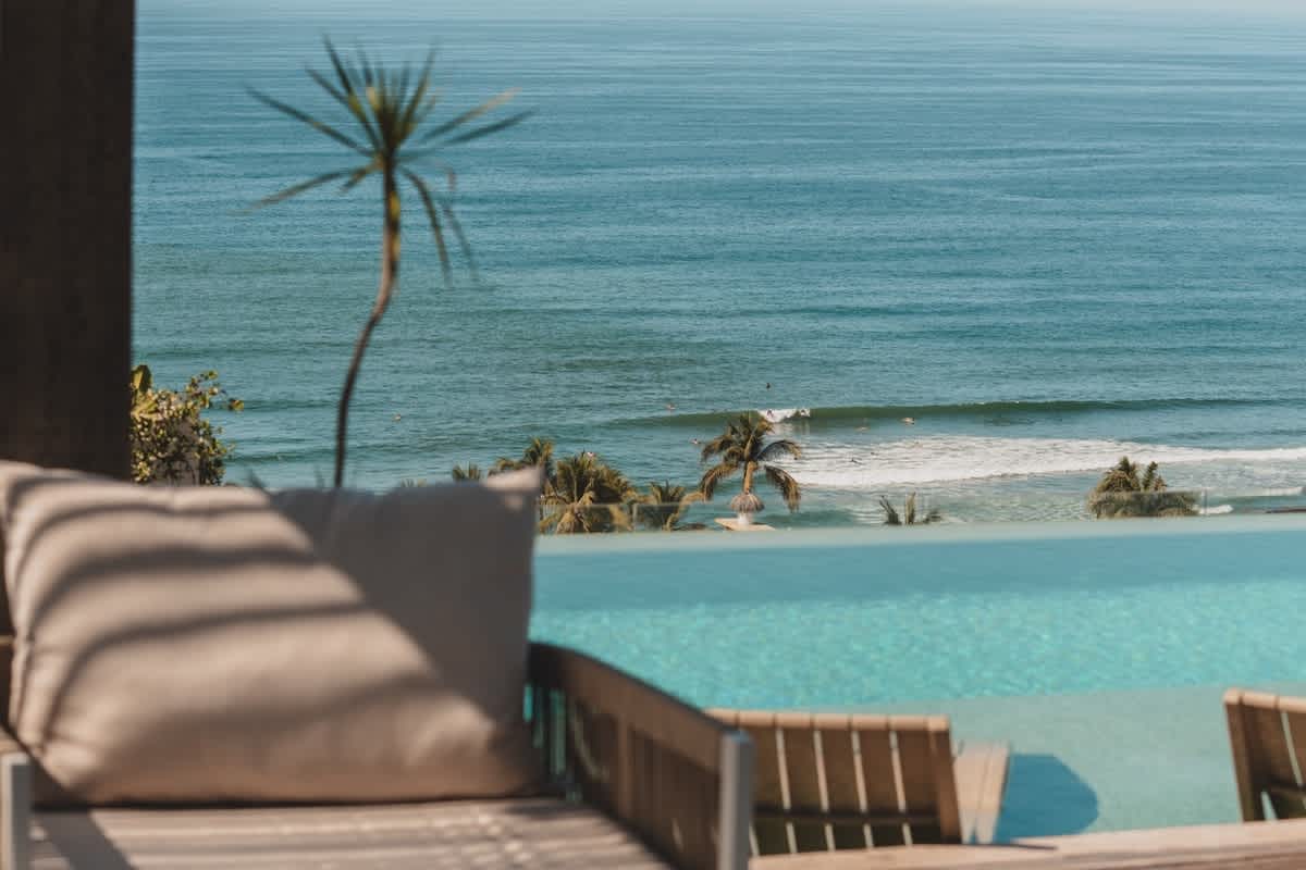 Close-up view of the infinity pool's edge, showcasing the calm blue ocean with distant surfers and a sandy beach, framed by a lounge chair with a neutral cushion and tropical foliage.