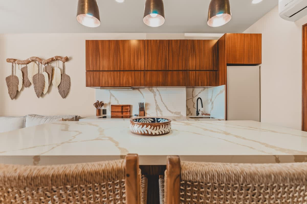 A modern kitchen with a large white quartz island and matching backsplash, featuring dark wood cabinetry and three bronze pendant lights overhead. A decorative bowl sits on the island, and a wall hanging with feather-like elements adorns the wall above the sofa in the background.