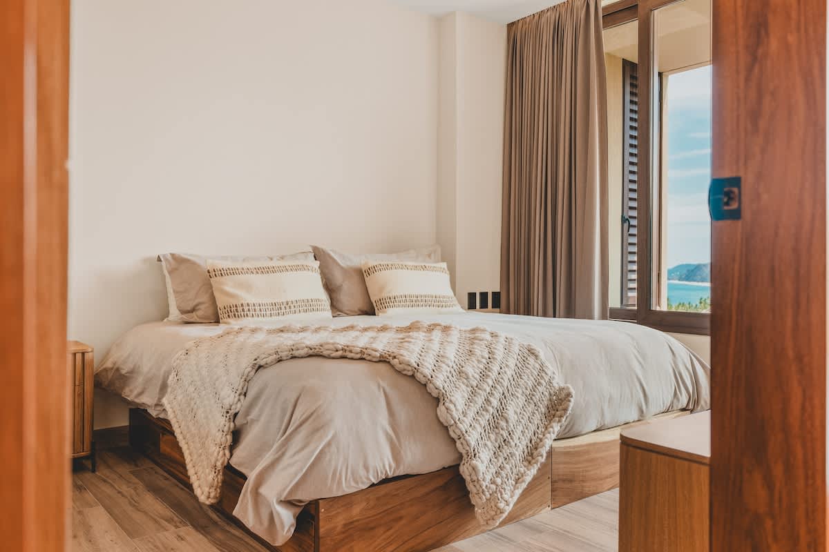 Cozy bedroom with a queen-sized bed featuring light grey bedding and a chunky knit throw, flanked by wooden nightstands, and a large window offering a partial ocean view.