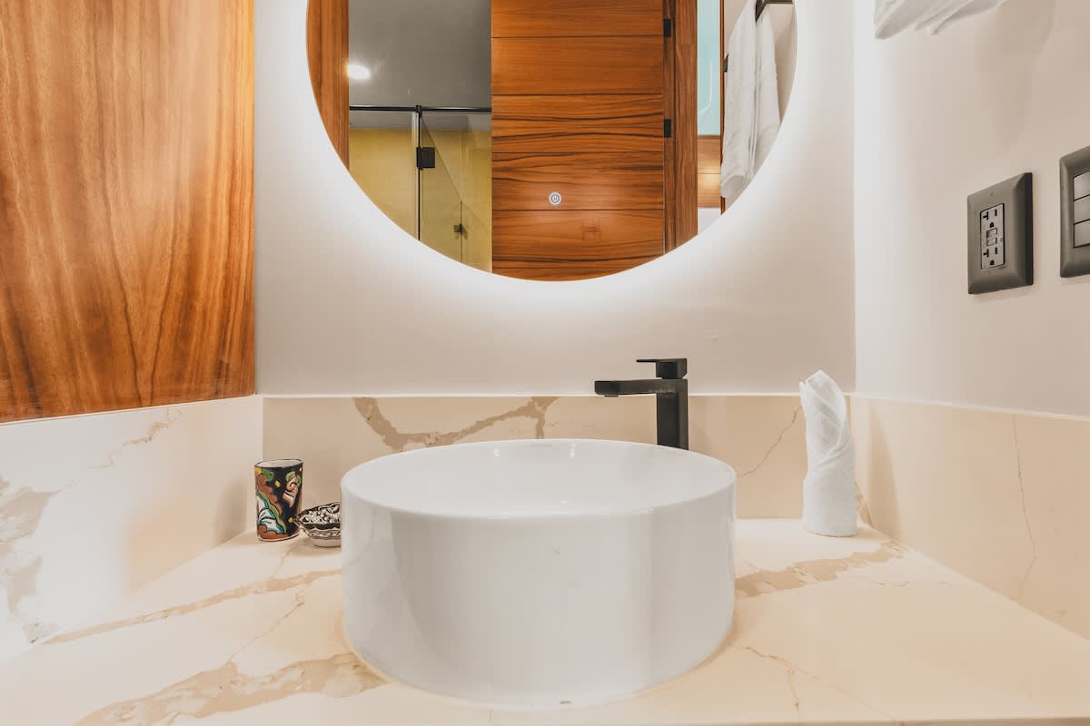 Detailed view of the modern bathroom vanity featuring a white vessel sink, a sleek black faucet, and a large round illuminated mirror above a marble-patterned countertop.
