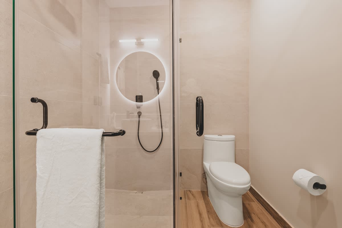Modern bathroom with a glass-enclosed shower featuring a rain showerhead and an illuminated mirror, alongside a white toilet and grab bars for accessibility.