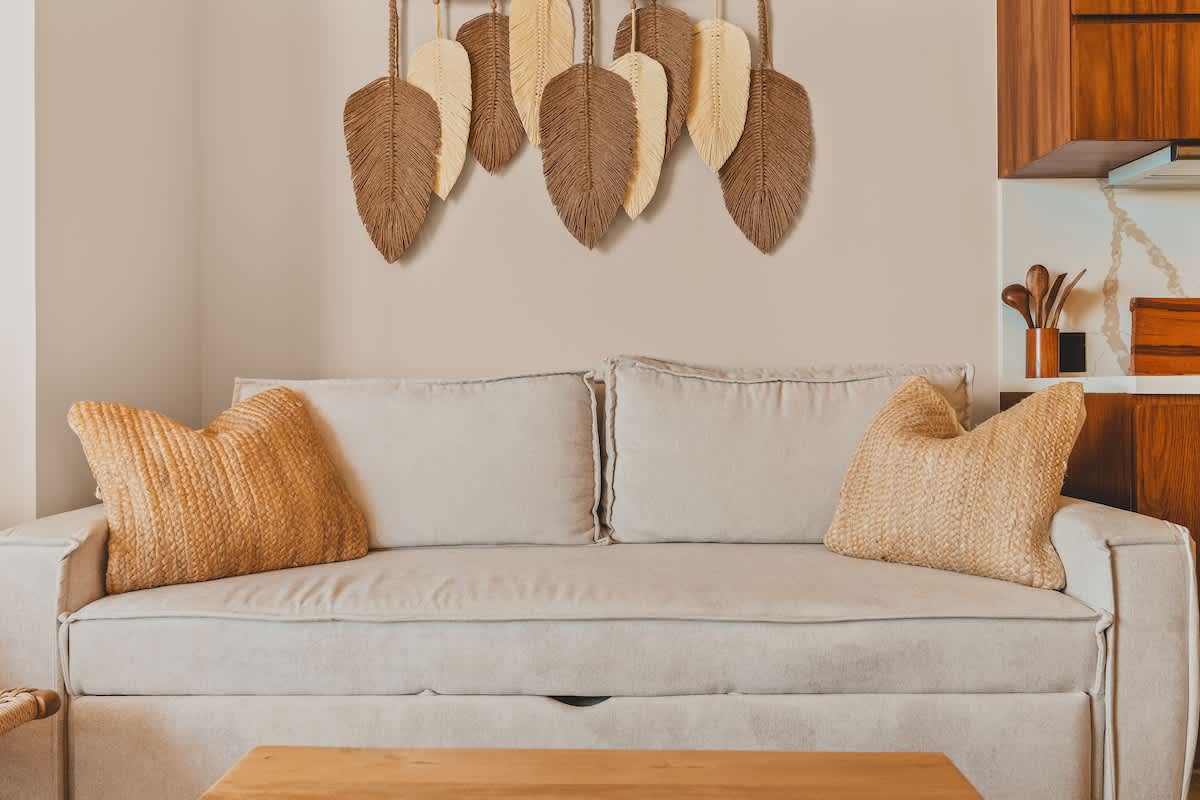 A comfortable light-colored sofa with two textured yellow throw pillows sits against a neutral wall, adorned with decorative woven leaf-shaped wall art in natural tones.