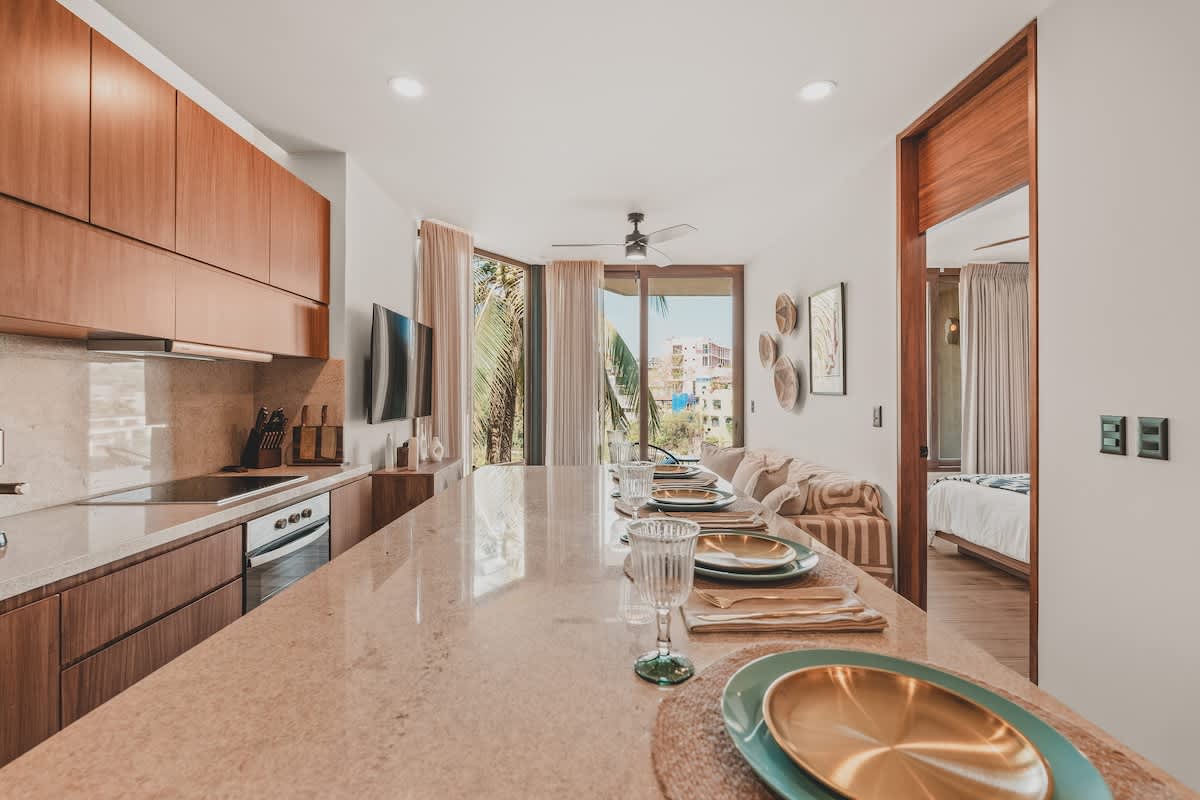 Open-concept dining and living area with a long stone island set for meals, offering views of the tropical outdoors through expansive windows and a glimpse into a bedroom.