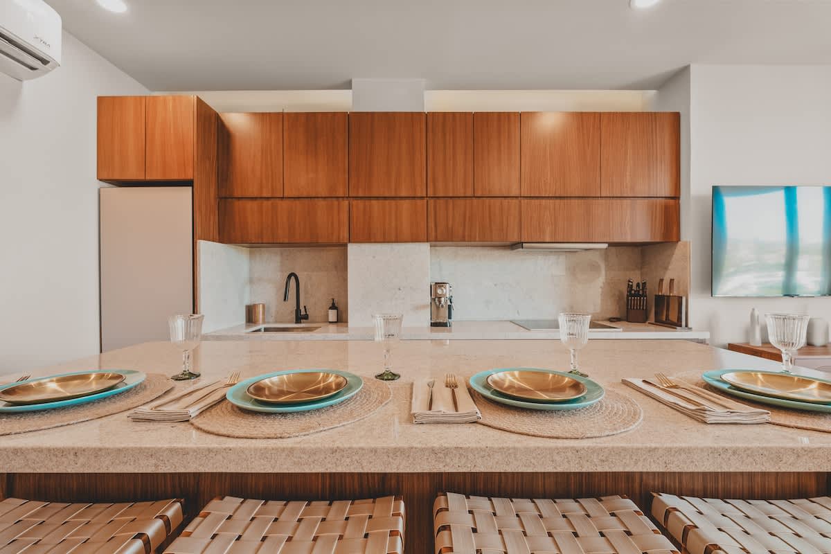 Modern kitchen island with multiple place settings and woven leather stools, backed by sleek wooden cabinetry, a built-in refrigerator, and an air conditioning unit.