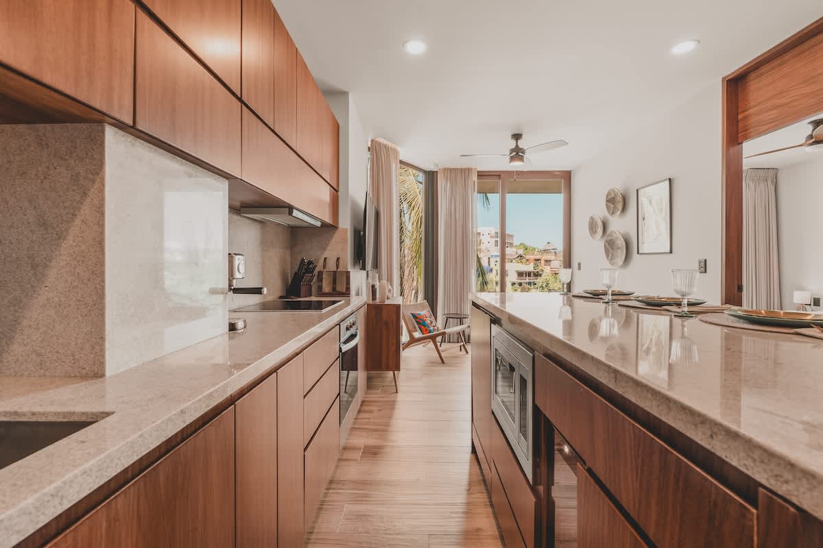 Expansive galley kitchen with rich wooden cabinetry and light stone countertops, equipped with a microwave, oven, stovetop, and espresso machine, leading to an open living area.