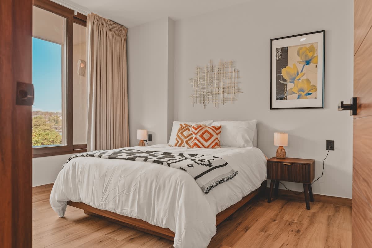 Bright bedroom featuring a queen-sized bed with crisp white bedding and vibrant orange patterned pillows, flanked by wooden nightstands with small lamps, and a large window with light curtains offering an outdoor view.