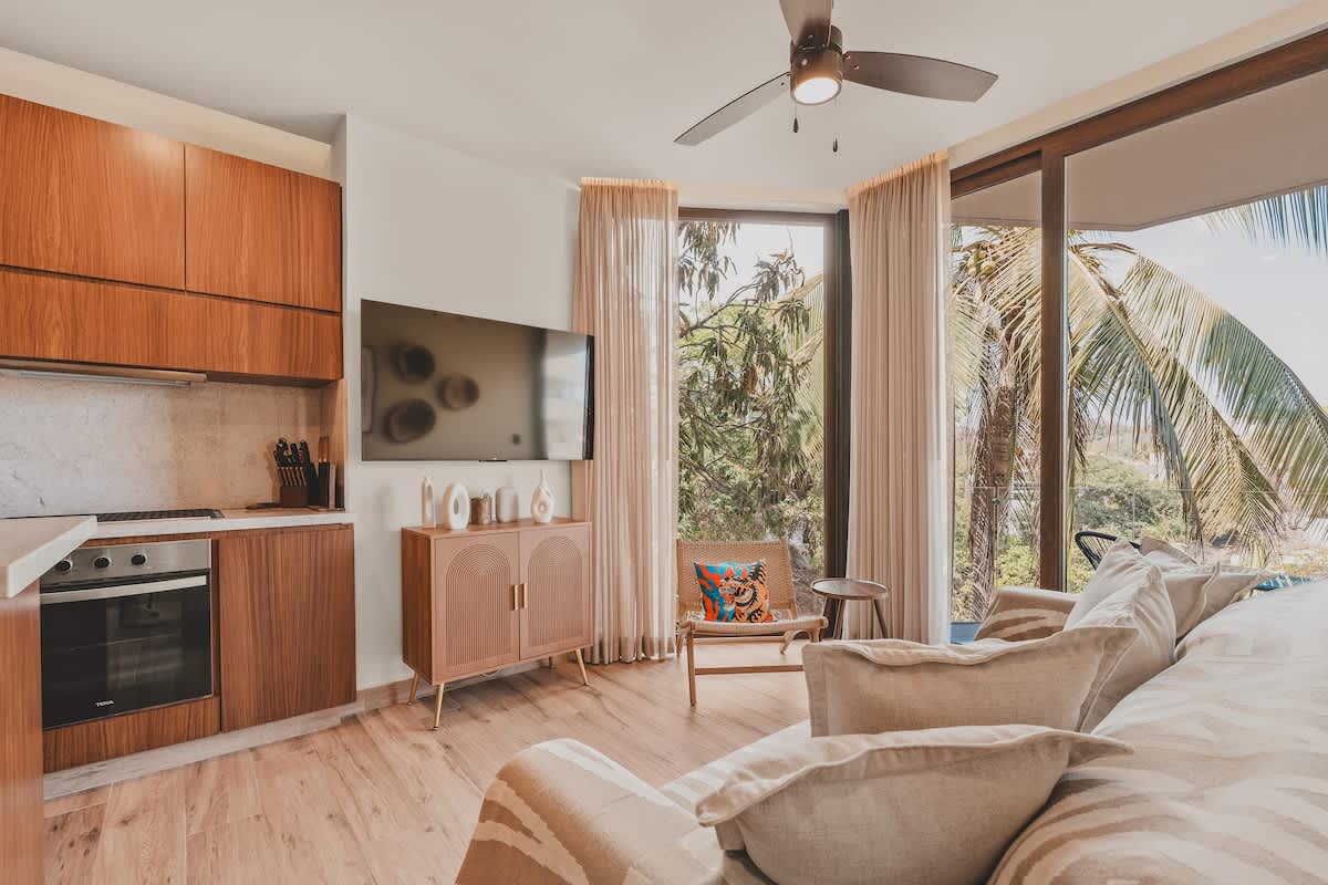 A modern living room with light wood floors, a beige sofa, and an integrated kitchen area with wooden cabinets and a built-in oven. Large windows and a sliding glass door reveal tropical palm trees and a bright outdoor scene.