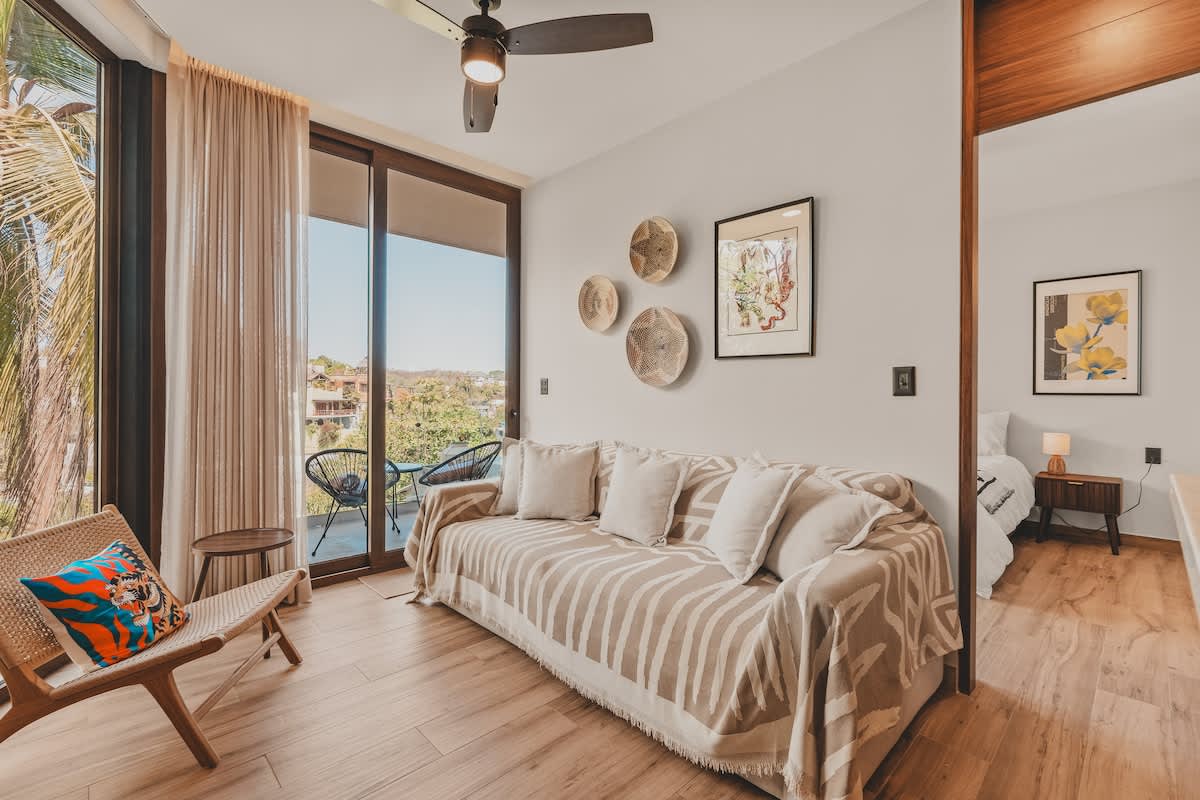 Cozy living room featuring a sofa with a patterned blanket and light pillows, a wooden accent chair, and large sliding glass doors opening to a balcony with outdoor seating and lush views.