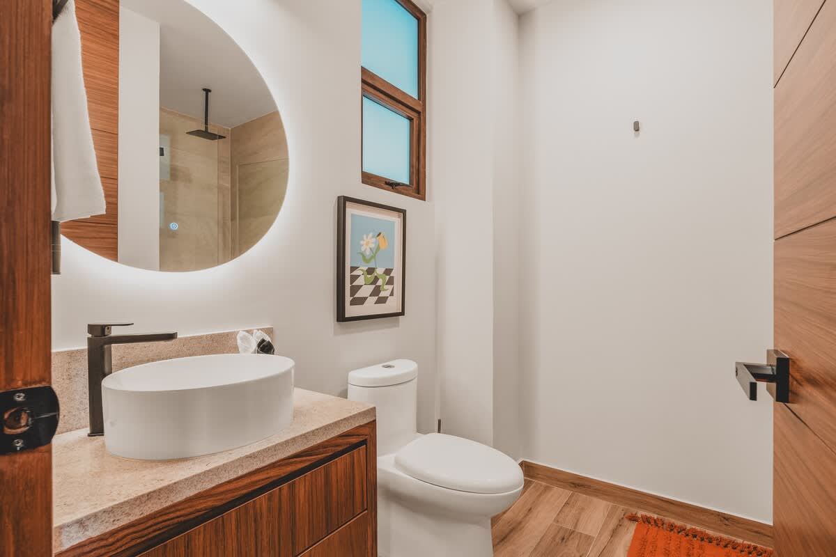 Modern bathroom featuring a white vessel sink on a wooden vanity, a toilet, and a large round backlit mirror, complemented by a small window and framed artwork.