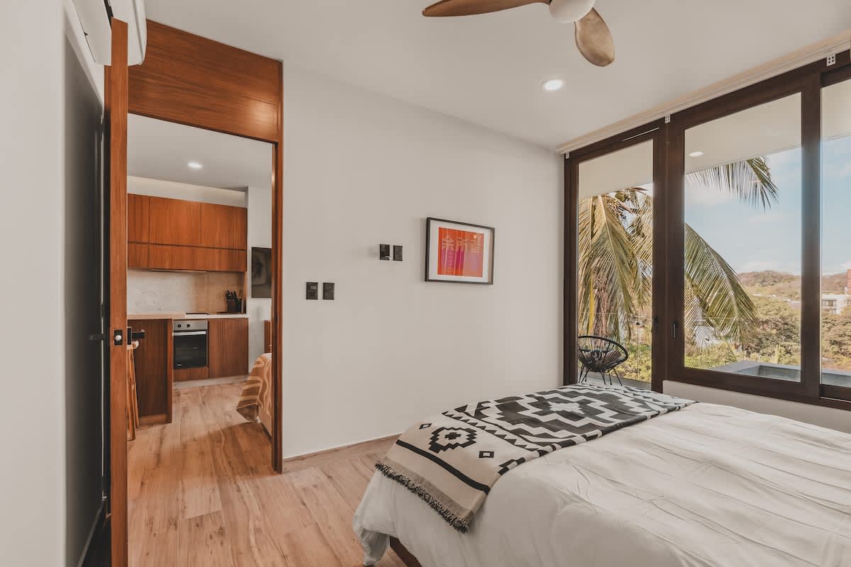 A bedroom with light wood floors and a queen bed, featuring a view of tropical foliage through large windows. A wooden doorway leads to a modern kitchen area, creating an open-concept feel.