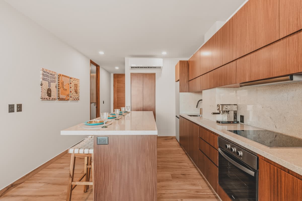 Modern kitchen featuring a long island with dining settings and a galley-style counter with integrated appliances, all framed by warm wooden cabinetry and light stone surfaces.