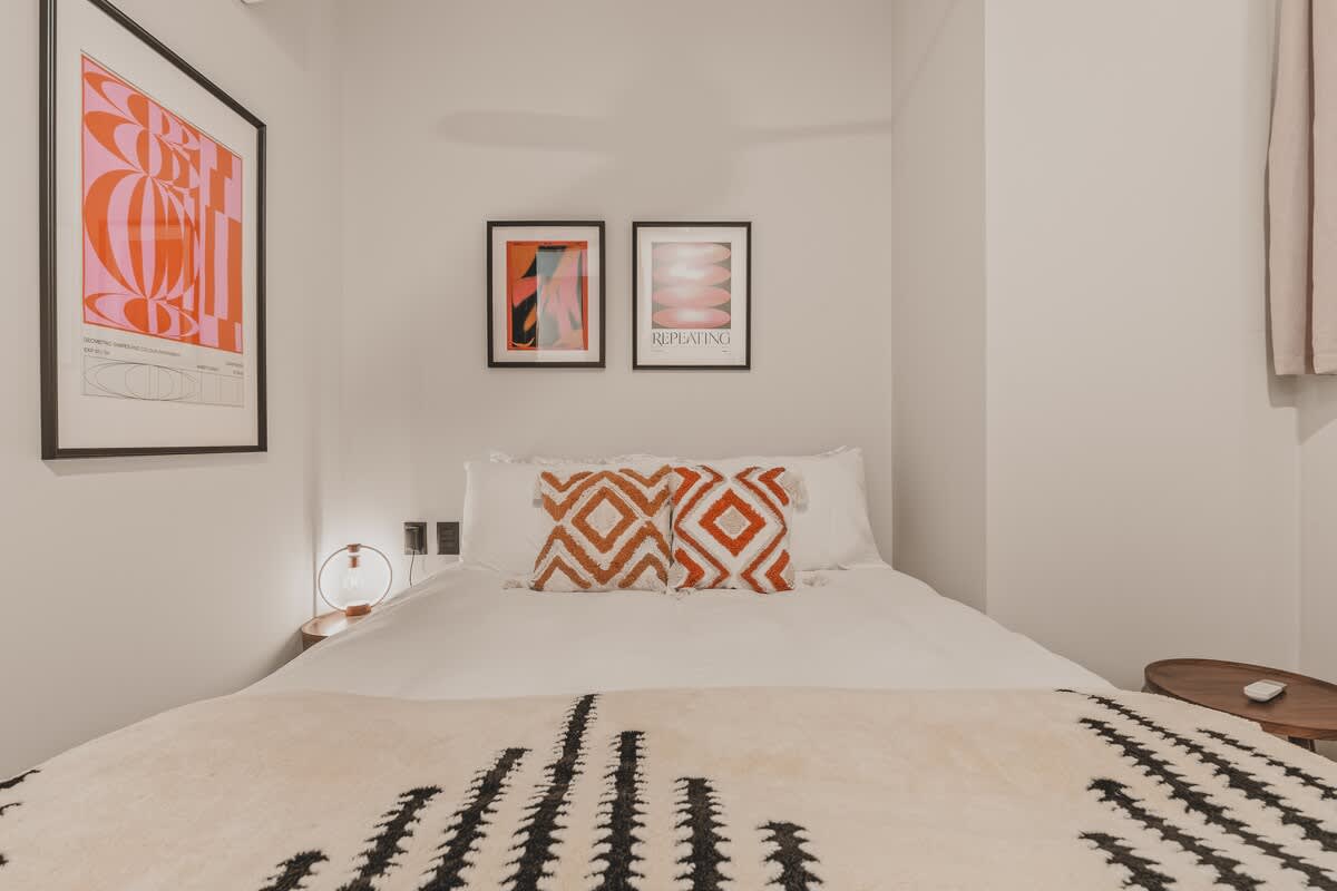 Close-up of a queen bed with white linens, two orange and white geometric patterned pillows, and a textured black and white throw blanket. Three framed abstract art pieces adorn the light gray wall above the bed.