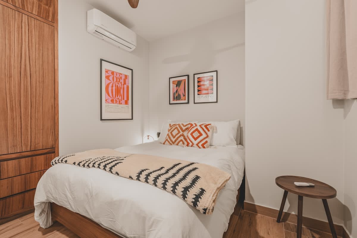 Modern bedroom with a queen bed, white bedding, and decorative orange and white pillows, complemented by a black and white patterned throw. A large wooden wardrobe is on the left, and an air conditioning unit is mounted above the bed.