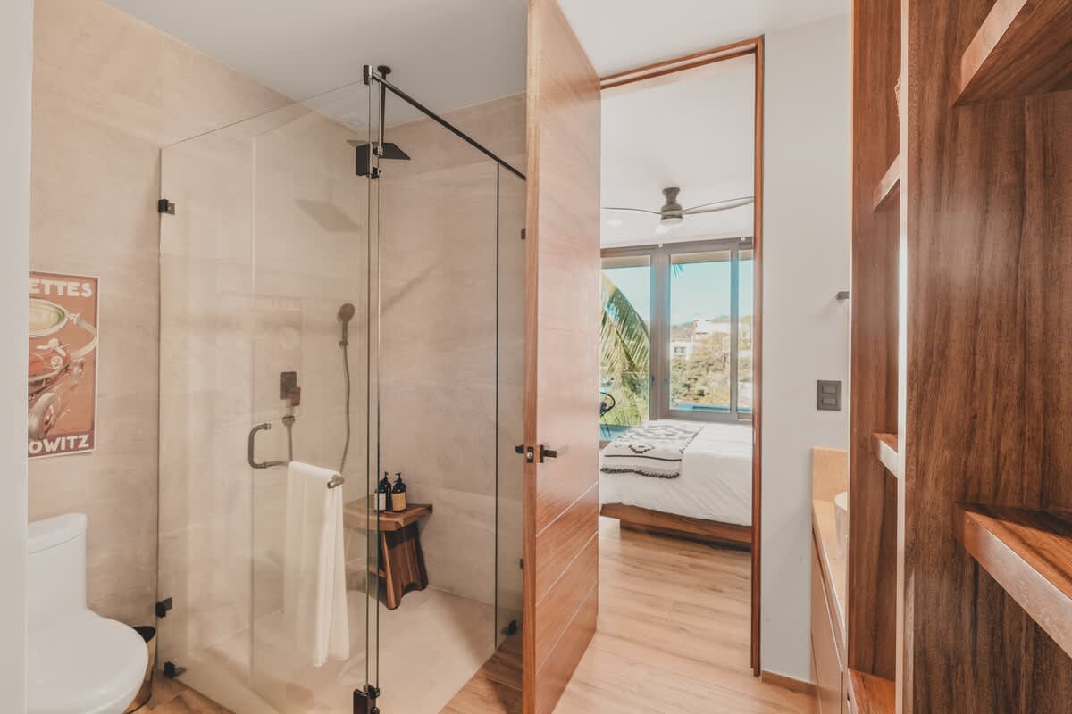 View from the bathroom showing the glass-enclosed shower and toilet, with a wooden door leading to the vanity area and a glimpse of the bedroom with a bed and large window.