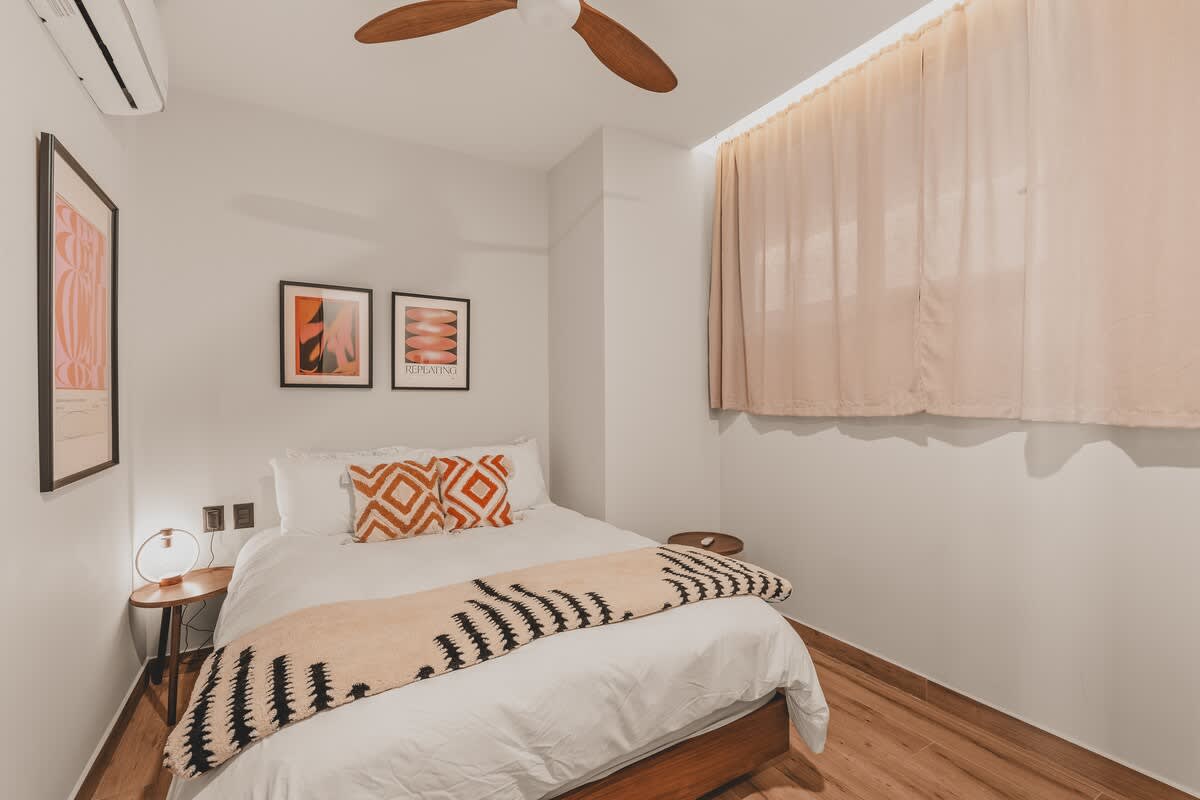 Cozy bedroom with a queen-sized bed featuring white bedding, orange patterned pillows, and a black and white patterned throw blanket. Two framed abstract art prints hang above the bed, and a wooden ceiling fan is visible.
