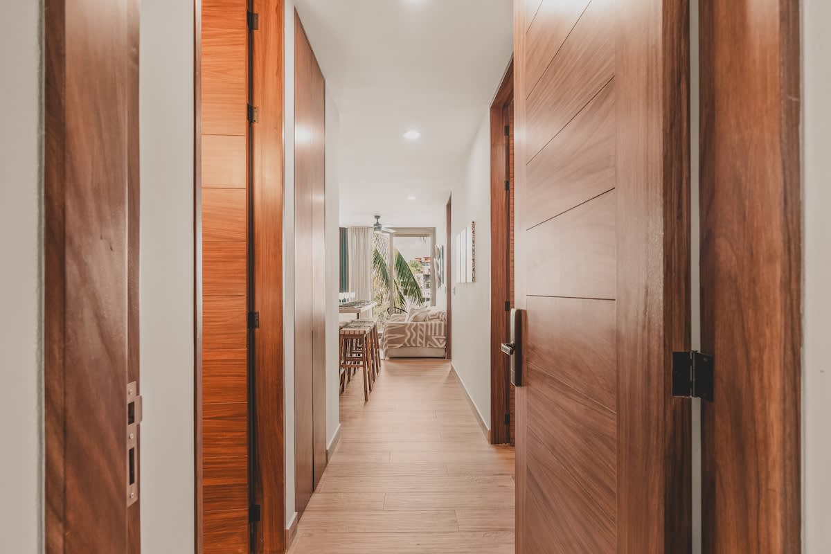 Bright hallway with light wood-look flooring and white walls, featuring multiple dark wood doors and door frames. The hallway leads towards a living area visible in the distance.