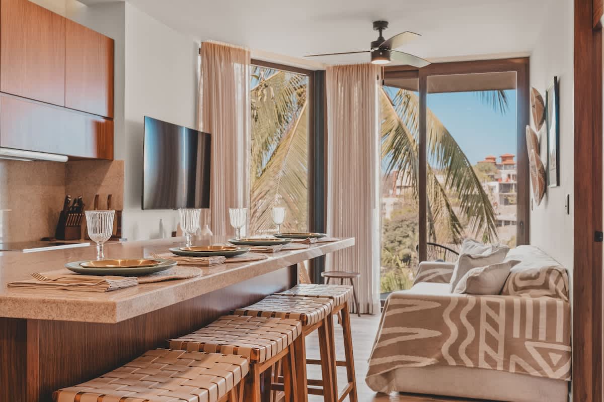Open-concept kitchen and dining area with a long island and bar stools, flowing into the living space with a sofa, all bathed in natural light from expansive windows offering views of palm trees and a balcony.
