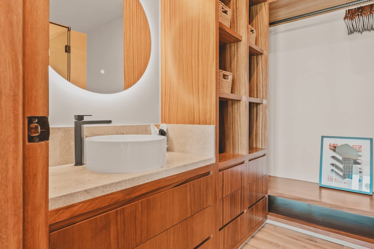 Stylish vanity area featuring a round backlit mirror above a white vessel sink with a black faucet, set into a light stone countertop, surrounded by rich wooden cabinetry and open shelving with woven baskets.