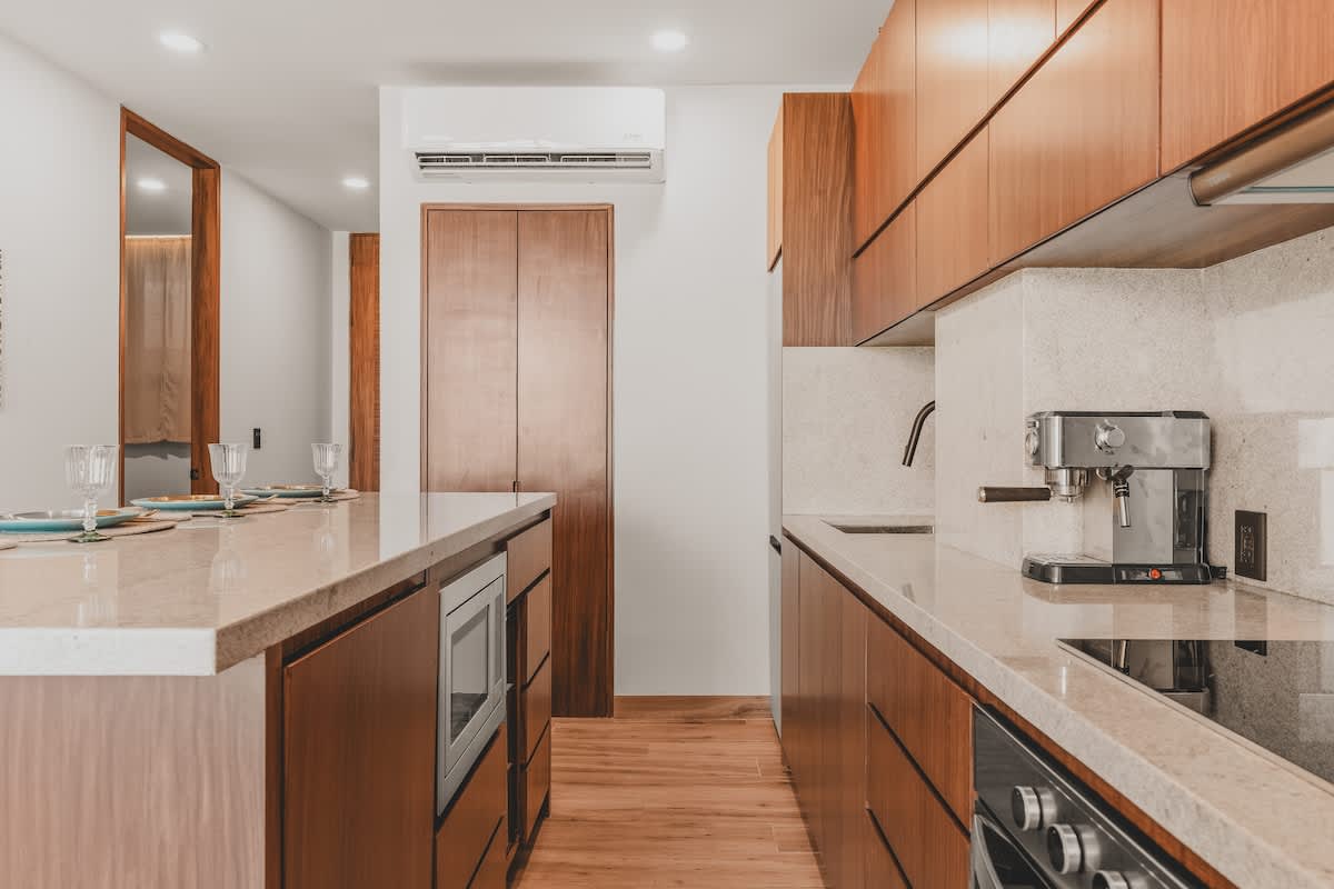 Galley kitchen with warm wooden cabinetry, light stone countertops, and integrated appliances including a microwave in the island and an oven, stovetop, and espresso machine along the main counter.
