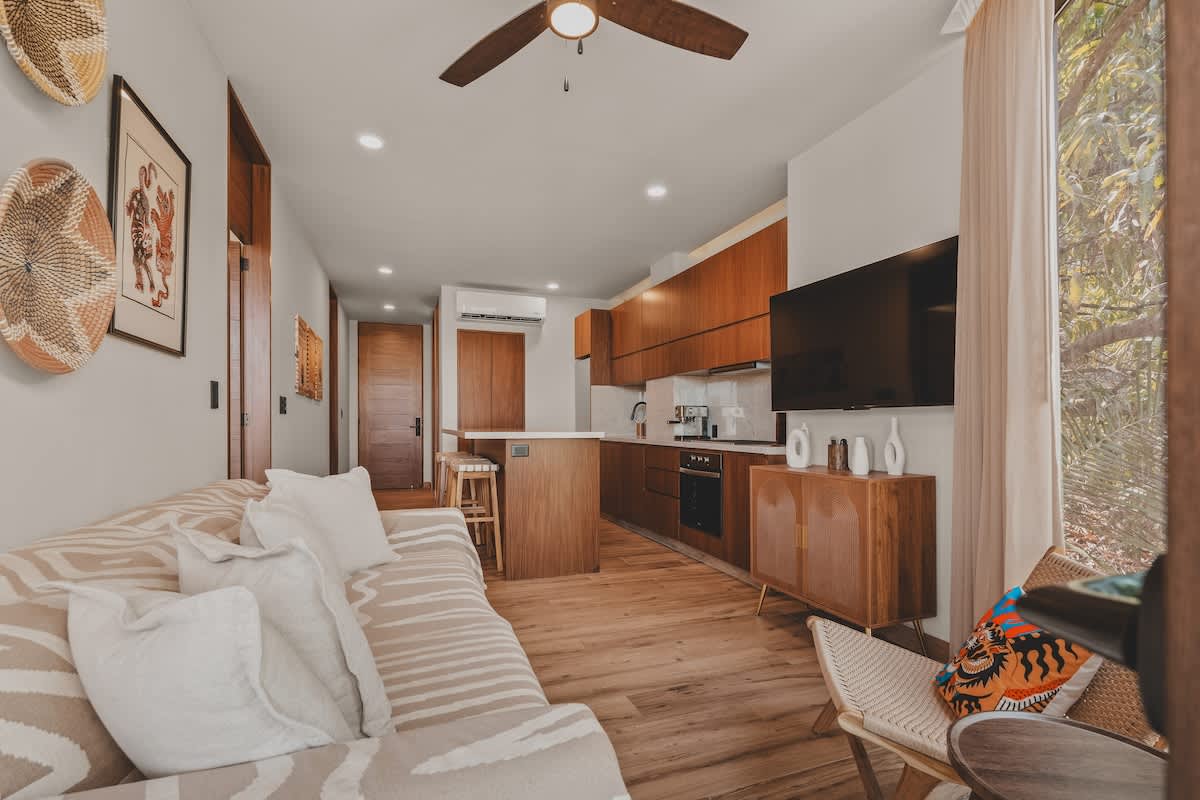 Modern open-concept living space featuring a light-colored sofa with patterned throw, a wall-mounted television, and a ceiling fan, flowing into a kitchen with warm wood cabinetry and light countertops.