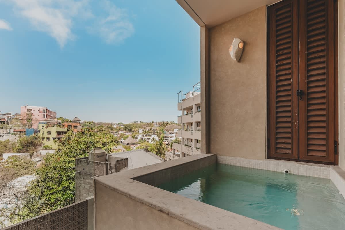 A private plunge pool on a balcony offers expansive views of the surrounding town and lush green hills under a clear blue sky. The pool is tiled and integrated into the balcony structure.