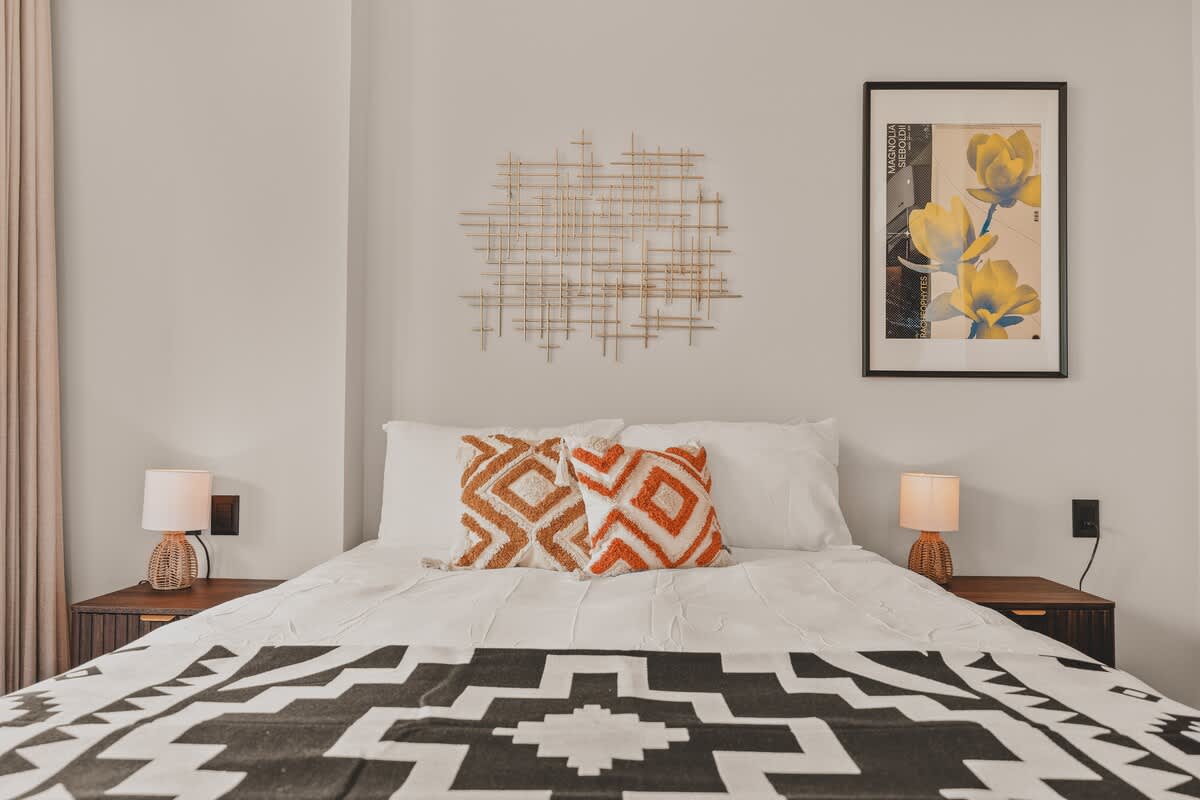 A close-up view of a queen bed in a bedroom, adorned with white bedding, two patterned throw pillows, and a black and white geometric blanket. A gold grid-like wall art piece is centered above the headboard.