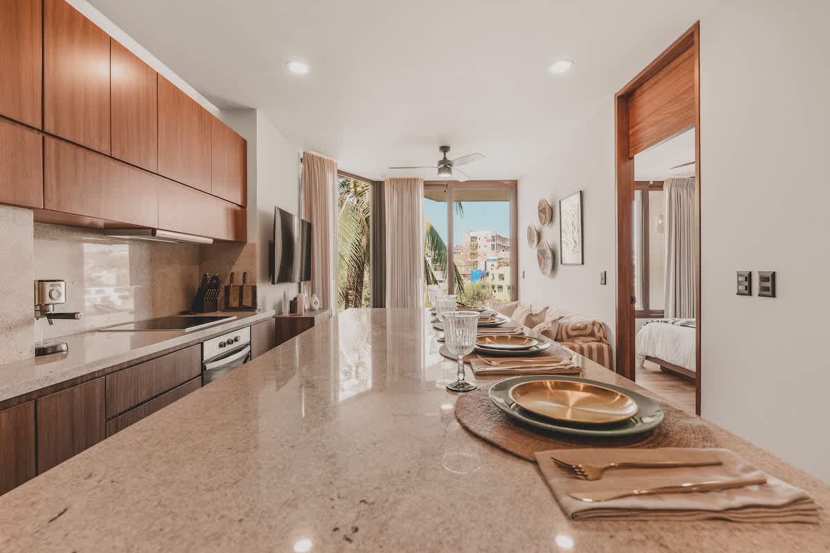 Bright dining area featuring a long, light-colored stone island with multiple place settings, overlooking a living space and large windows with a tropical outdoor view.