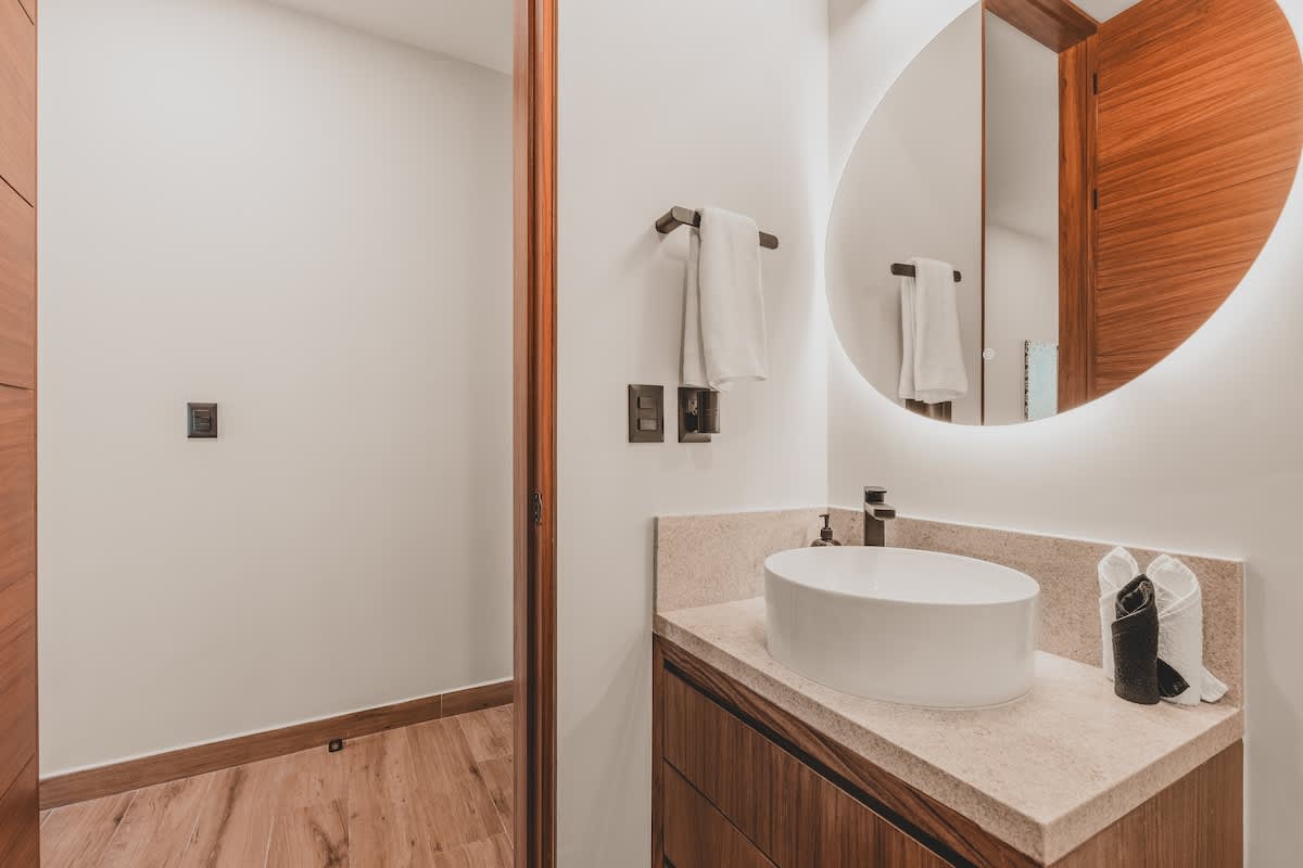Modern bathroom vanity featuring a white oval vessel sink on a light speckled countertop, with a large backlit oval mirror and dark wood cabinetry below. A towel rack with white towels is mounted on the wall.