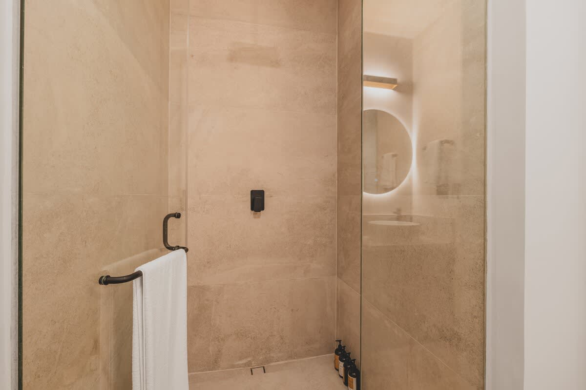 Contemporary shower stall with light beige stone-look tiles, a glass enclosure, and a dark metal towel bar holding a white towel. Three dark bottles of toiletries are visible on a shelf inside.