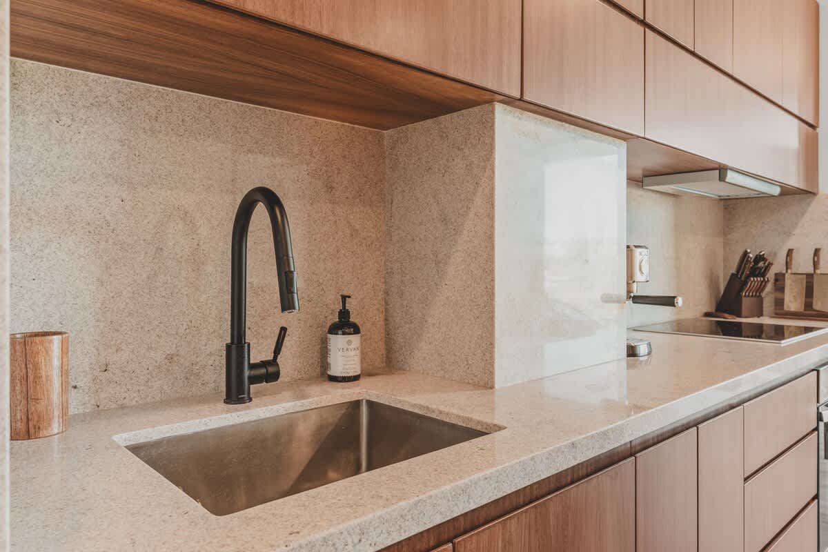 Close-up of a contemporary kitchen sink with a matte black gooseneck faucet and soap dispenser, set into a light stone countertop with a matching backsplash.