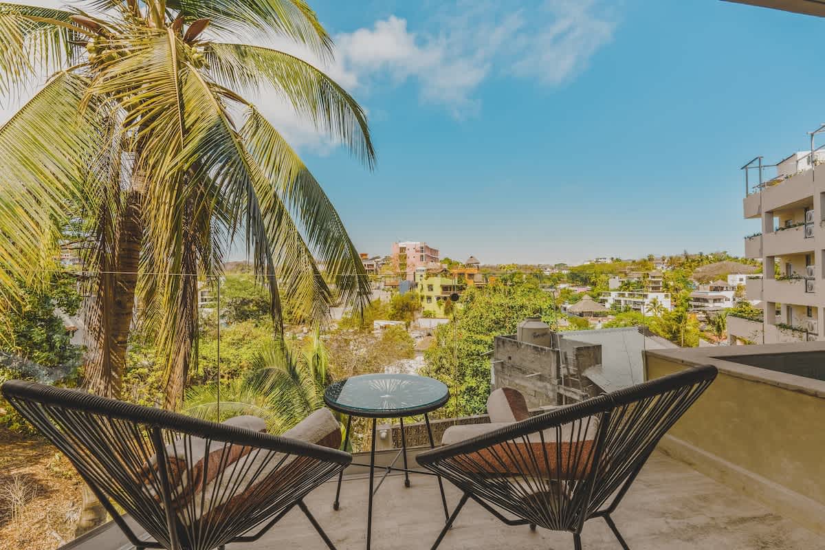 A private balcony features two stylish black string chairs and a small round table, providing a serene spot with a view of lush palm trees and the charming rooftops of a distant town.