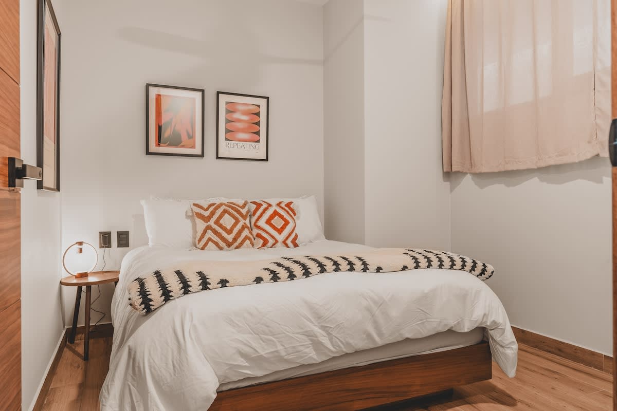 Serene bedroom featuring a queen bed with white bedding and decorative pillows, a patterned throw, and a small round wooden nightstand with a lamp. A window with light-colored curtains is visible on the right.