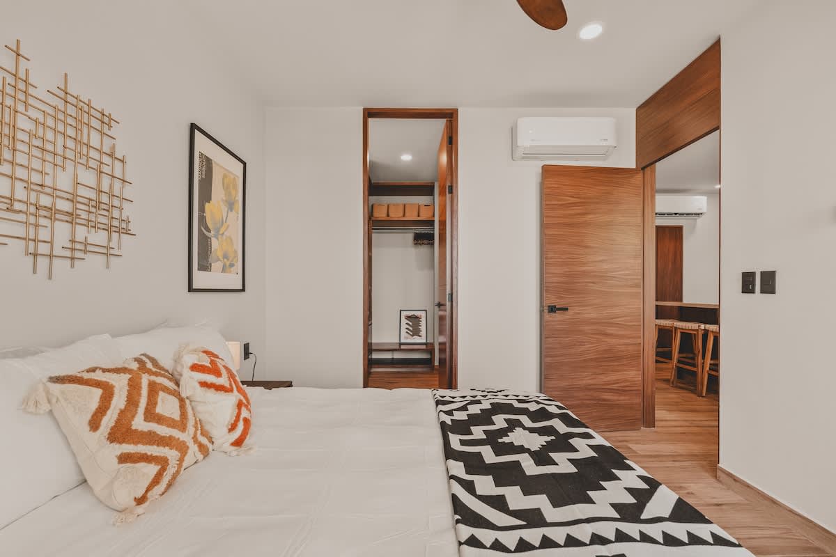View of the bedroom from a different angle, showcasing the bed with patterned pillows and a black and white geometric blanket, a decorative metal wall art piece, and an open doorway leading to a closet with built-in shelving.